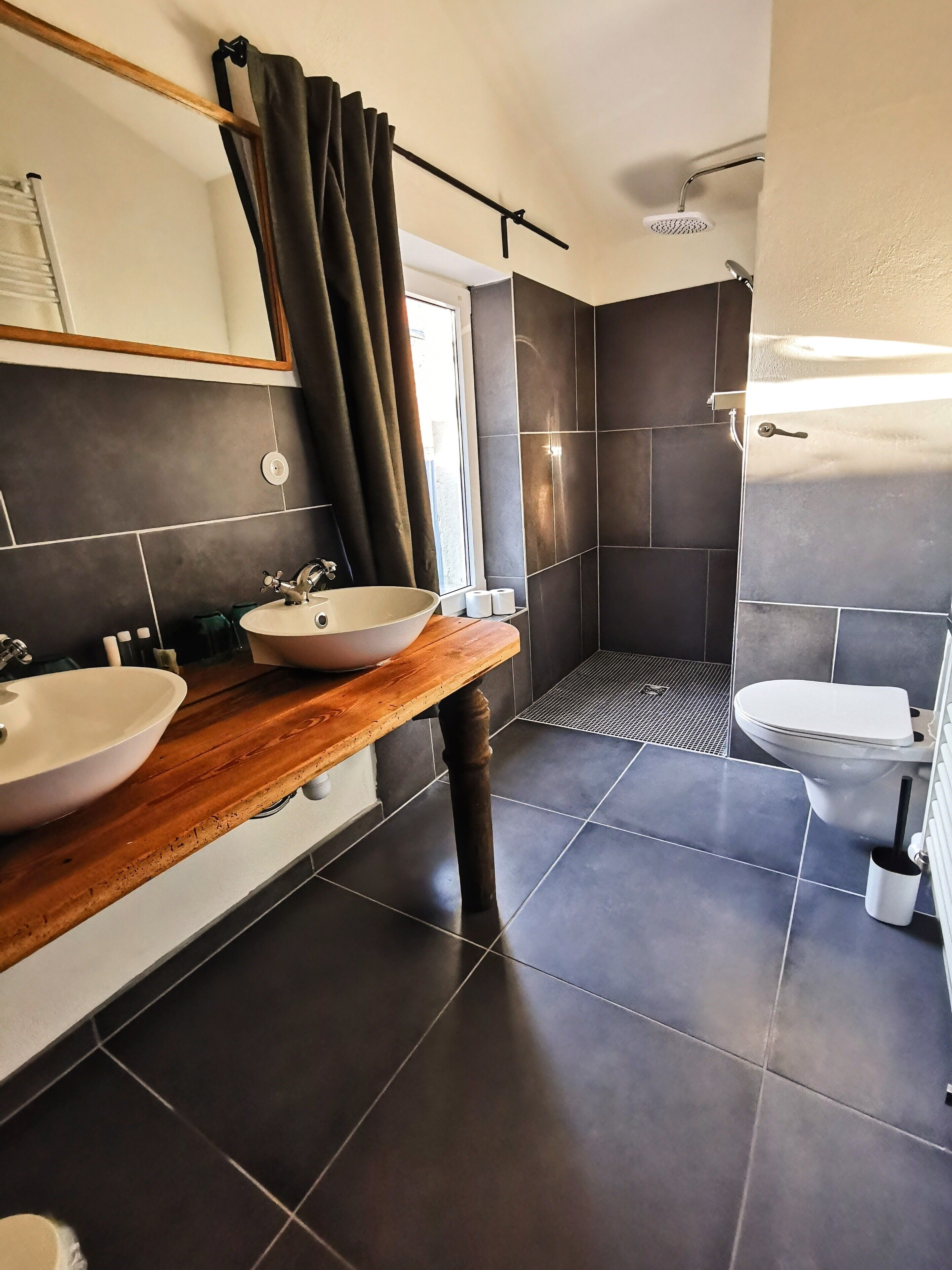 Un baño moderno con azulejos oscuros, tocador de madera, dos lavamanos, ducha e inodoro.