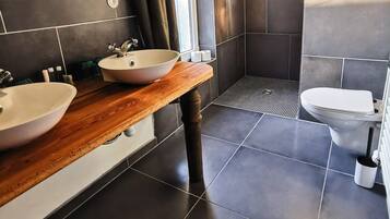 Un baño moderno con azulejos oscuros, tocador de madera, dos lavamanos, ducha e inodoro.