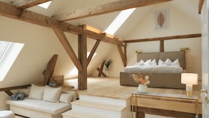 Un dormitorio acogedor en el ático con vigas de madera a la vista, una cama con ropa blanca, un pequeño sofá y una mesita de noche con lámpara.