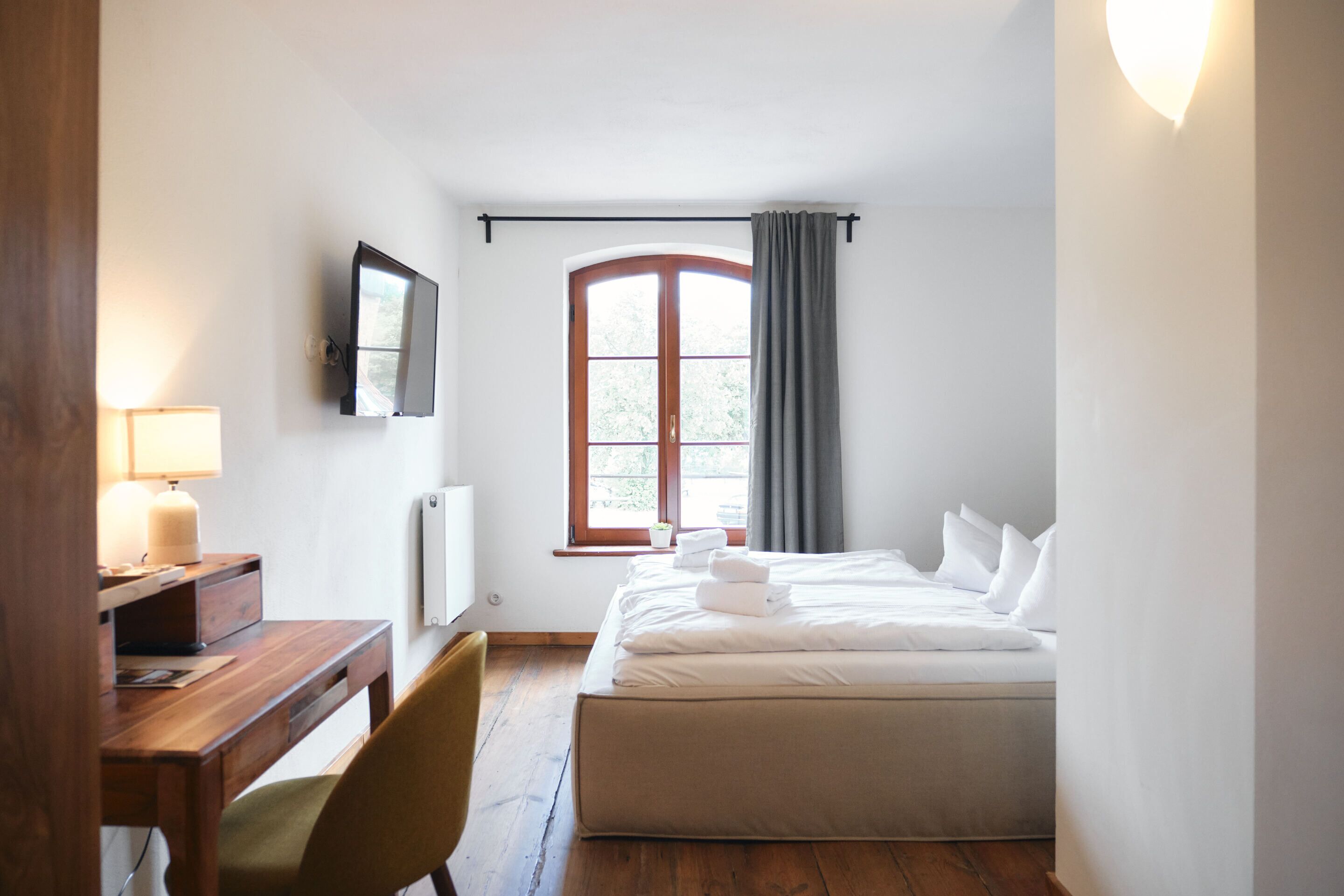 Ein Schlafzimmer mit Bett, Schreibtisch, Stuhl, einem Fenster mit Vorhängen und einem an der Wand montierten Fernseher.
