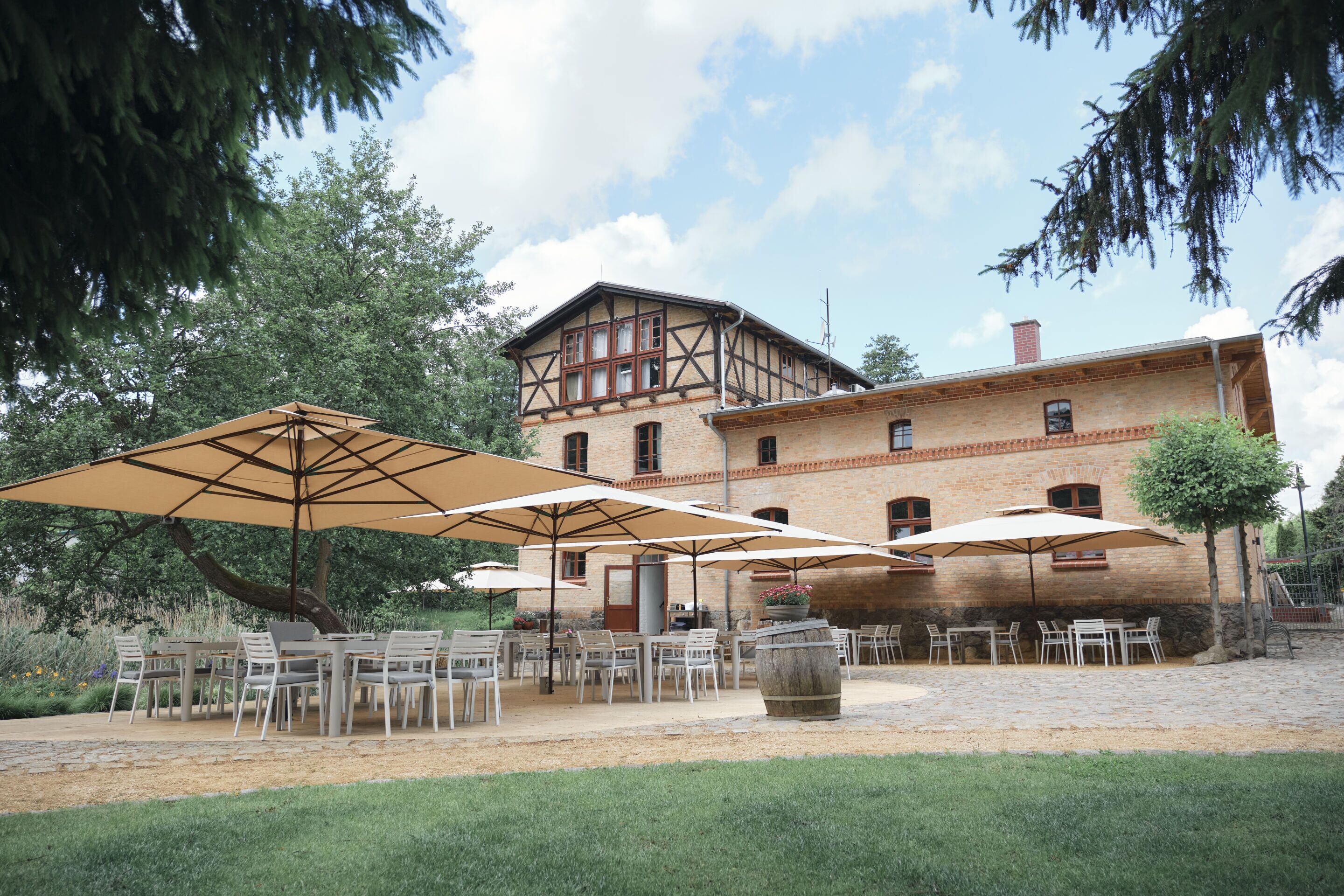 An outdoor patio with tables and chairs under umbrellas, a stone pathway, and a large building with a brick facade and wooden accents.
