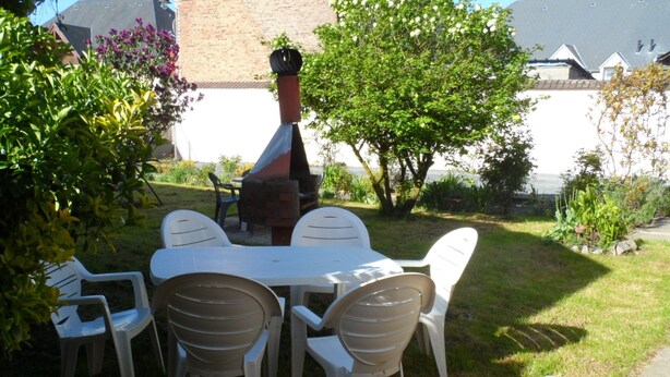 En udendørs terrasse med et bord og stole, omgivet af grøn vegetation og en murstensovn.