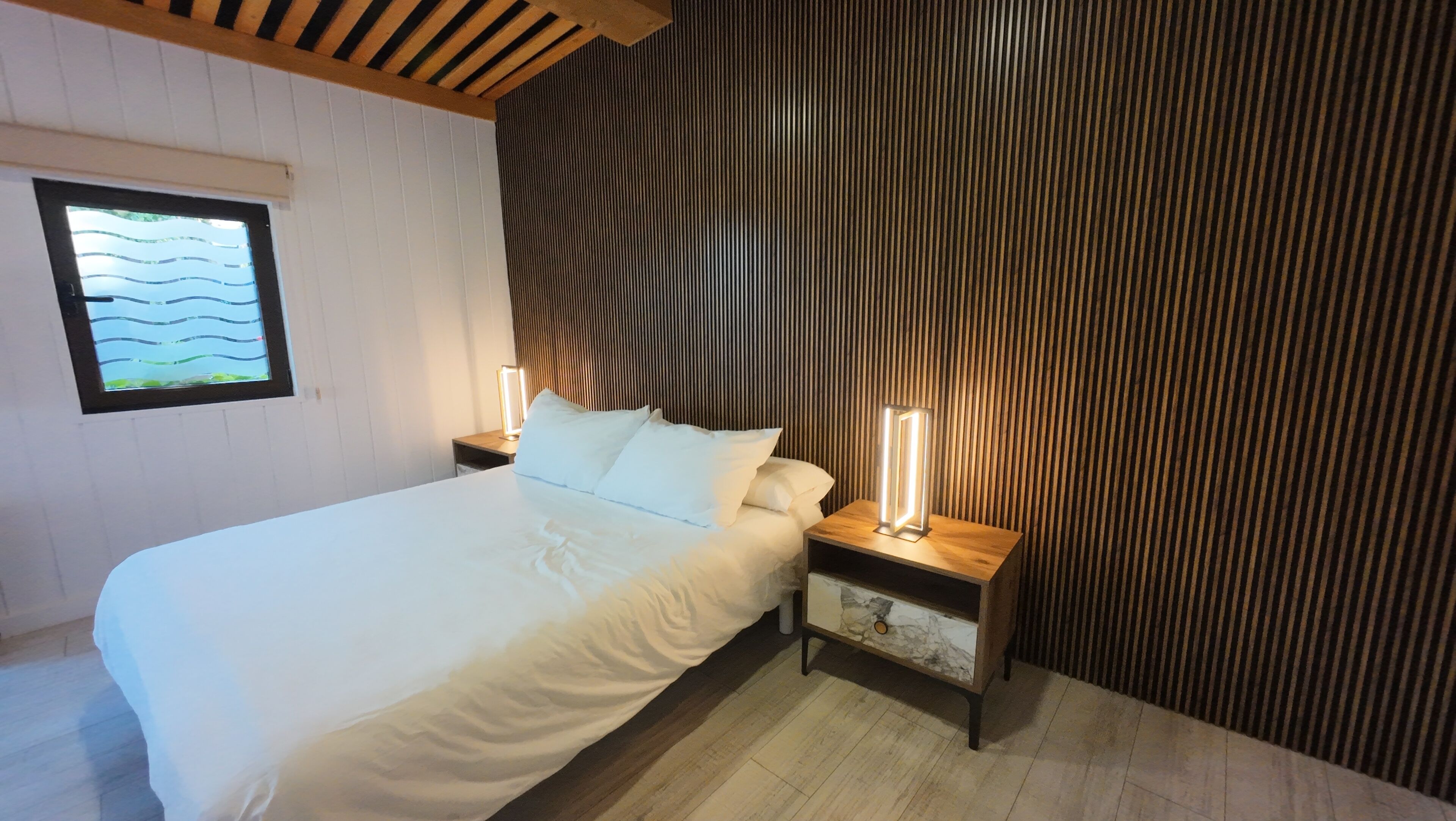 Un dormitorio con cama, dos mesitas de noche, una lámpara de noche, una ventana con vistas y una pared de listones de madera.