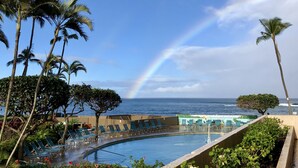 A pool area with lounge chairs, palm trees, and a view of the ocean with a rainbow.