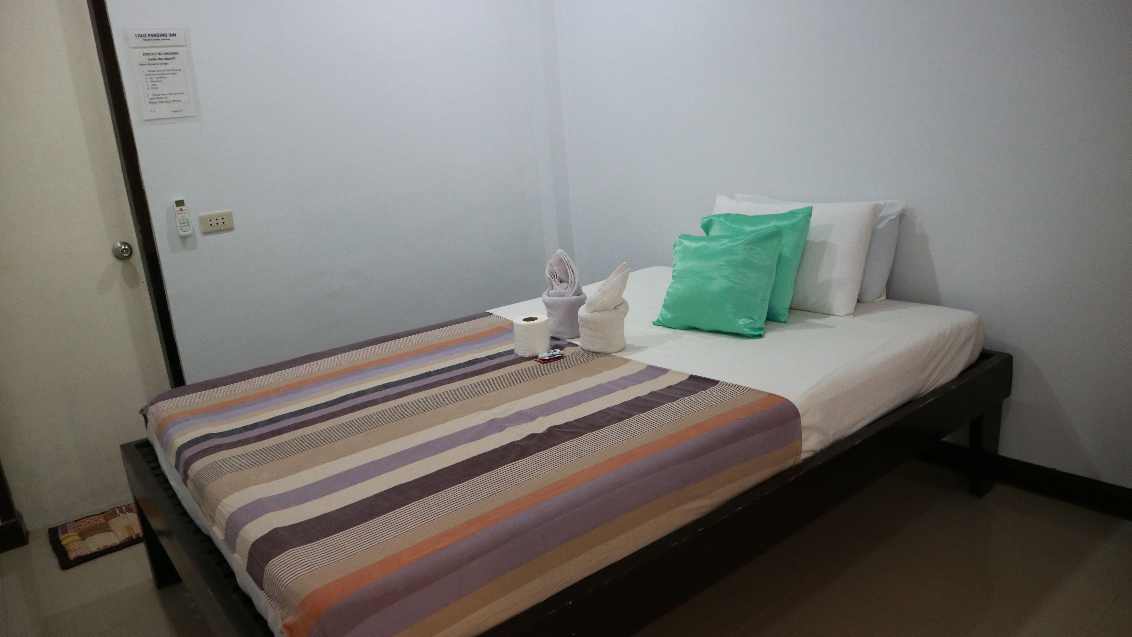 Un lit simple avec une couvre-lit à rayures, un oreiller vert et un oreiller blanc. Il y a une serviette et une télécommande sur le lit.