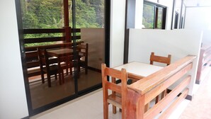 Una habitación con una mesa y sillas de madera, un banco y un amplio ventanal corredera que da a un balcón con vistas a la vegetación.