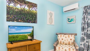 A room with a television displaying a beach scene, a wicker dresser, a wooden chair with a seashore pattern, and a wall-mounted air conditioner. - Prime Location. Hidden Gem. Steps to Beach. Boutique Bungalow. Pet Friendly.  (Juno Beach)