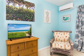 A room with a television displaying a beach scene, a wicker dresser, a wooden chair with a seashore pattern, and a wall-mounted air conditioner.