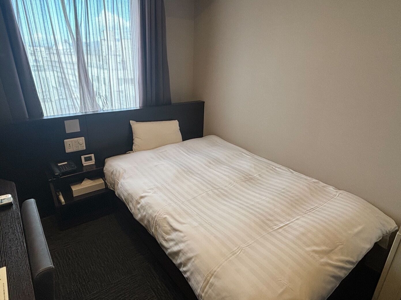 Ein Hotelzimmer mit Bett, Schreibtisch, Stuhl, einem Fenster mit Vorhängen und einer Wandbedieneinheit.