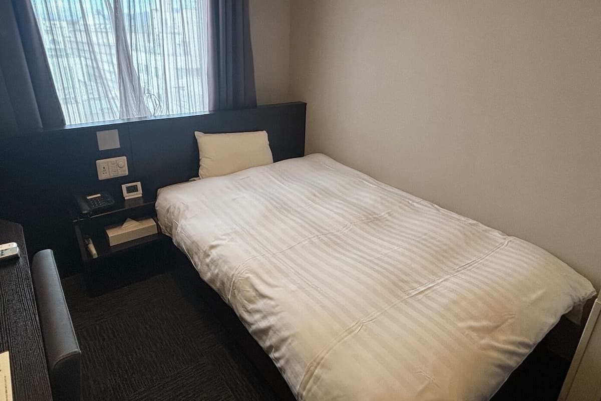 A hotel room with a bed, a desk, a chair, a window with curtains, and a wall-mounted control panel.