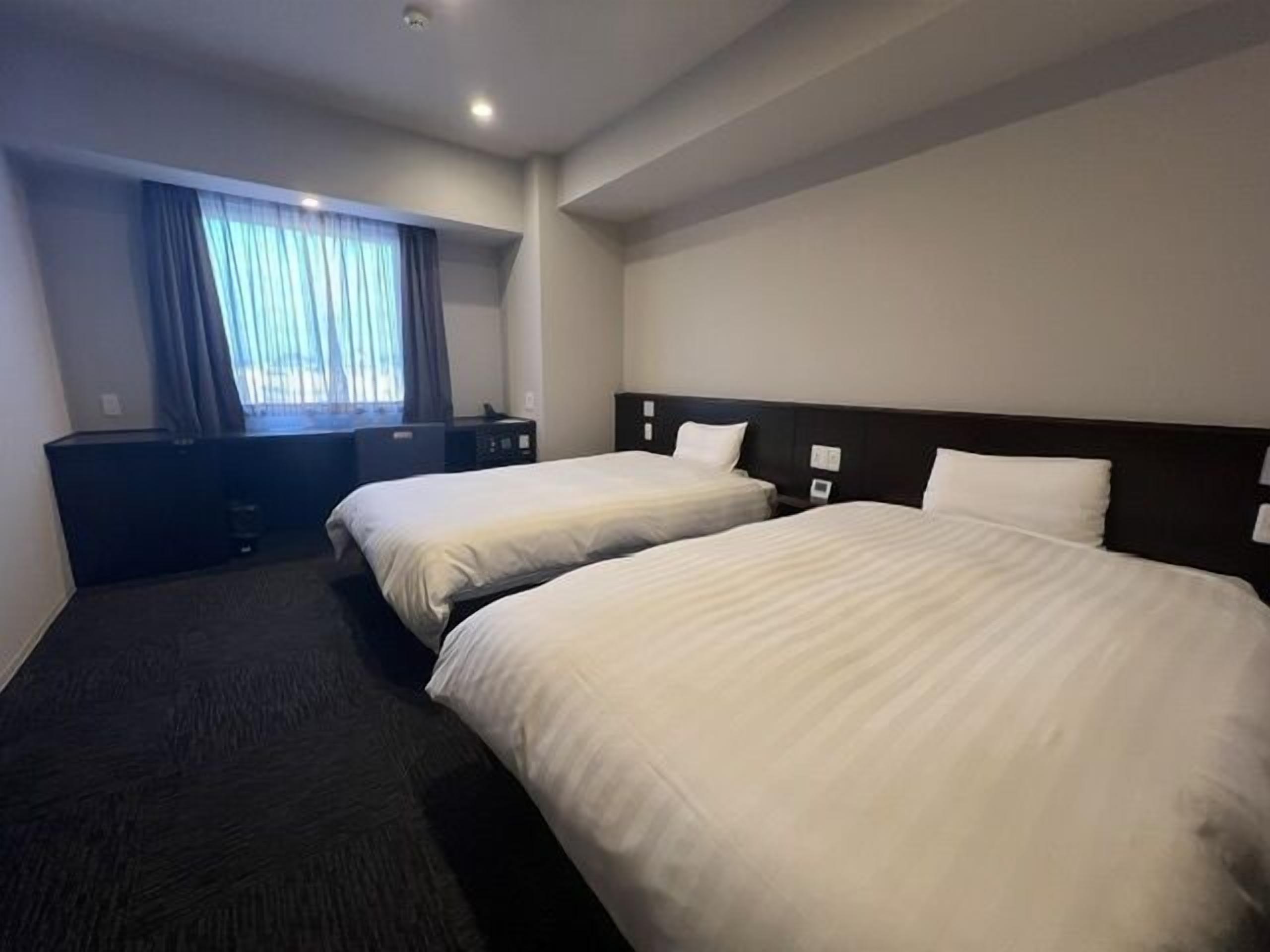 Ein Hotelzimmer mit zwei Betten, einem Schreibtisch, einem Stuhl, einem Fenster mit Vorhängen und einem Flachbildfernseher.