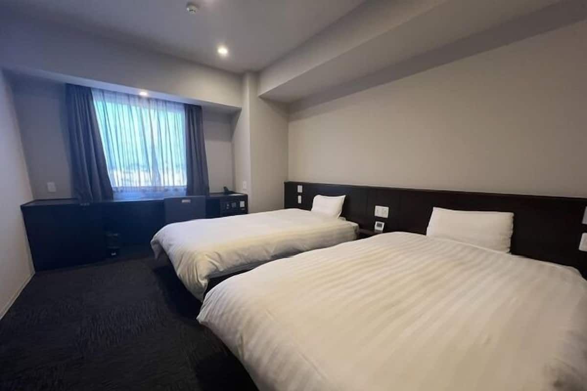 A hotel room with two beds, a desk, a chair, a window with curtains, and a flat-screen TV.