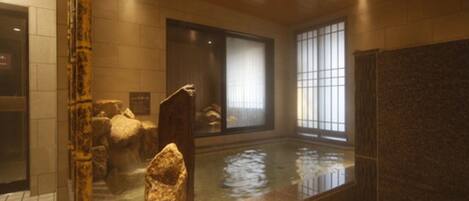 A traditional Japanese onsen with a wooden interior, a stone bath, and a view of the outside through a large window.