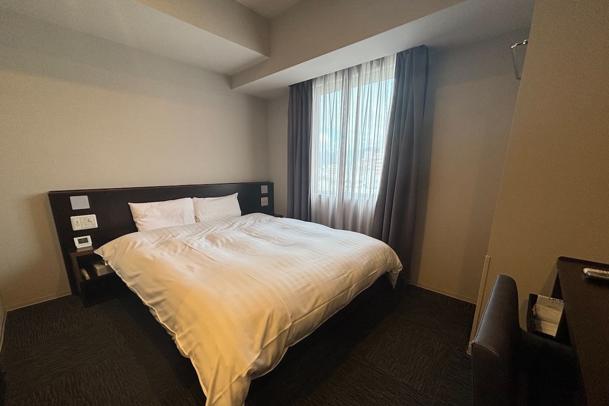A hotel room with a large bed, a bedside table with a lamp, and a window with curtains.