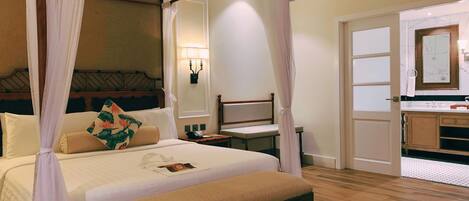 A hotel room with a bed, a bedside table, a chair, a bathroom with a mirror, and a wooden floor.