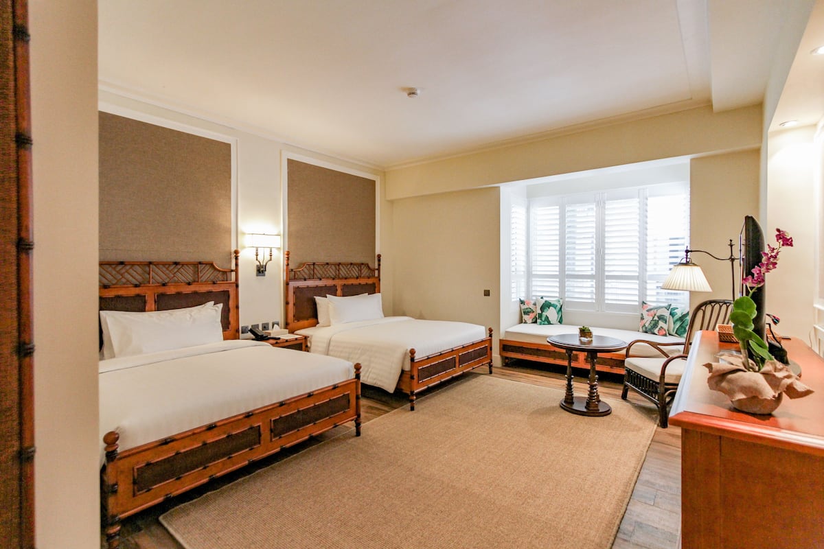 A hotel room with two wooden beds, a small table, a chair, and a window with blinds.