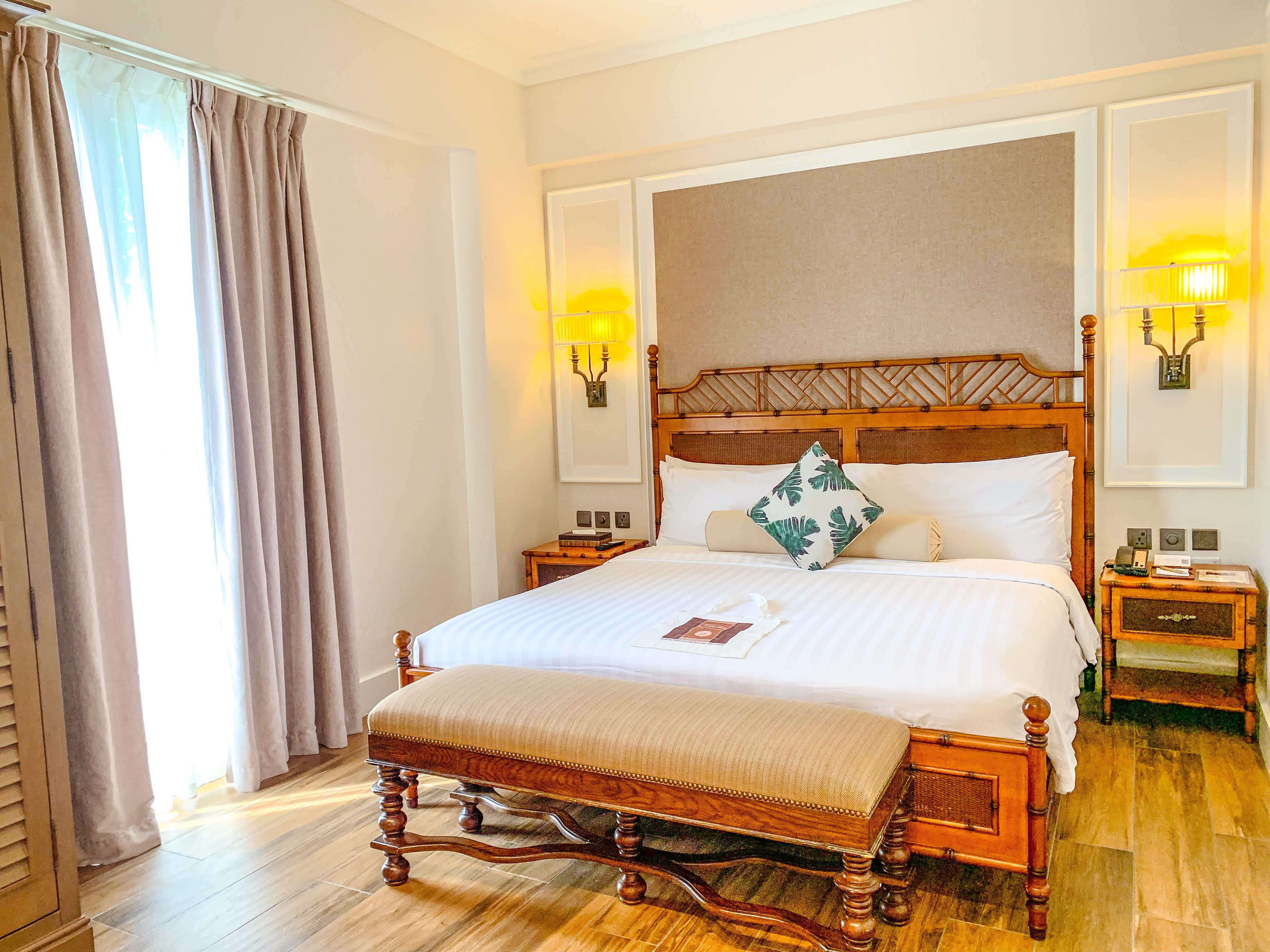 A hotel room with a large bed, wooden headboard, two bedside tables, a nightstand, and a window with curtains.