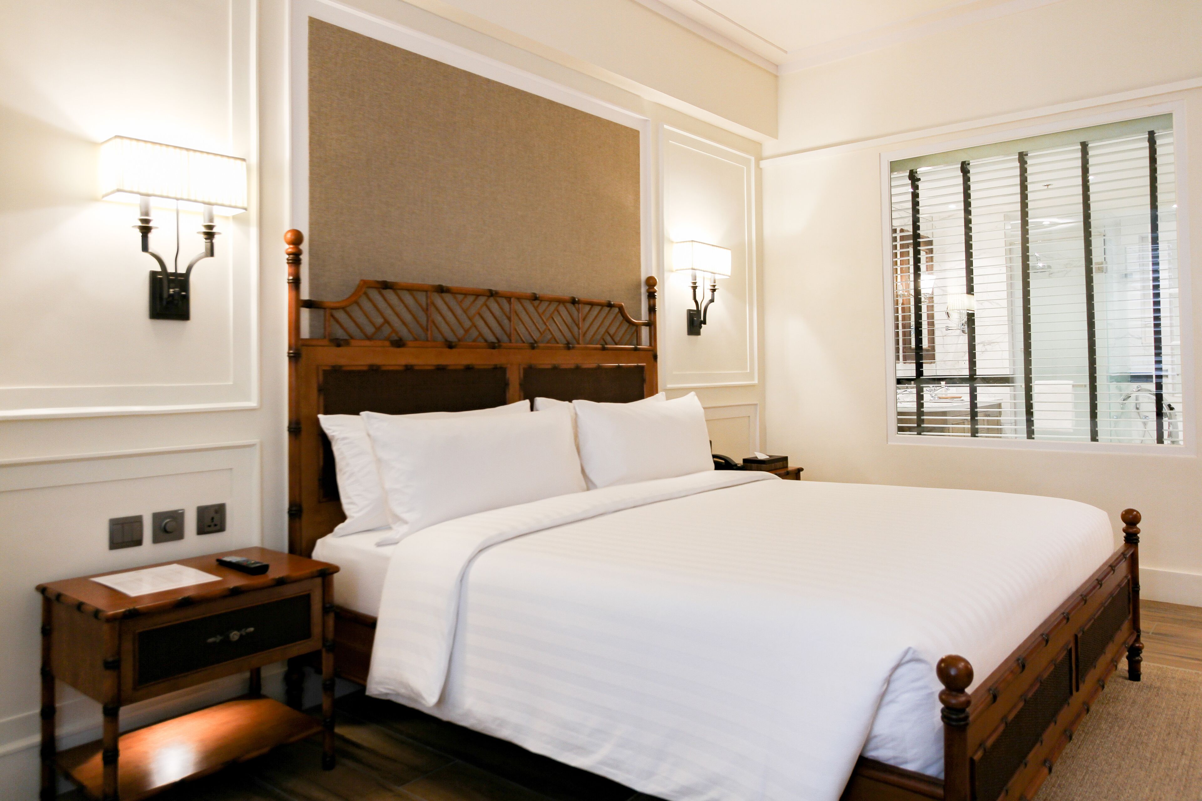 A neatly made bed with a wooden headboard, a bedside table with a lamp, and a window with blinds.