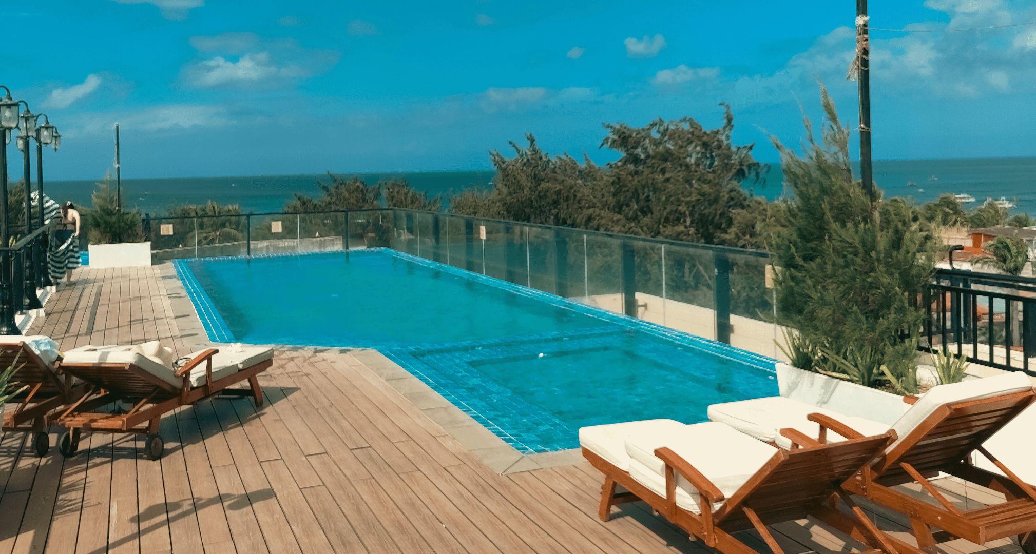 A rectangular swimming pool with a glass barrier, surrounded by wooden deck and lounge chairs.