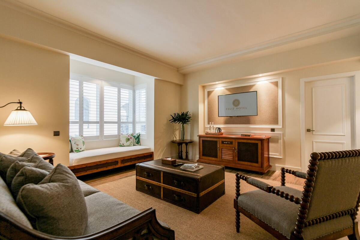 A living room with a sofa, a wooden coffee table, a television mounted on the wall, and a window with blinds.