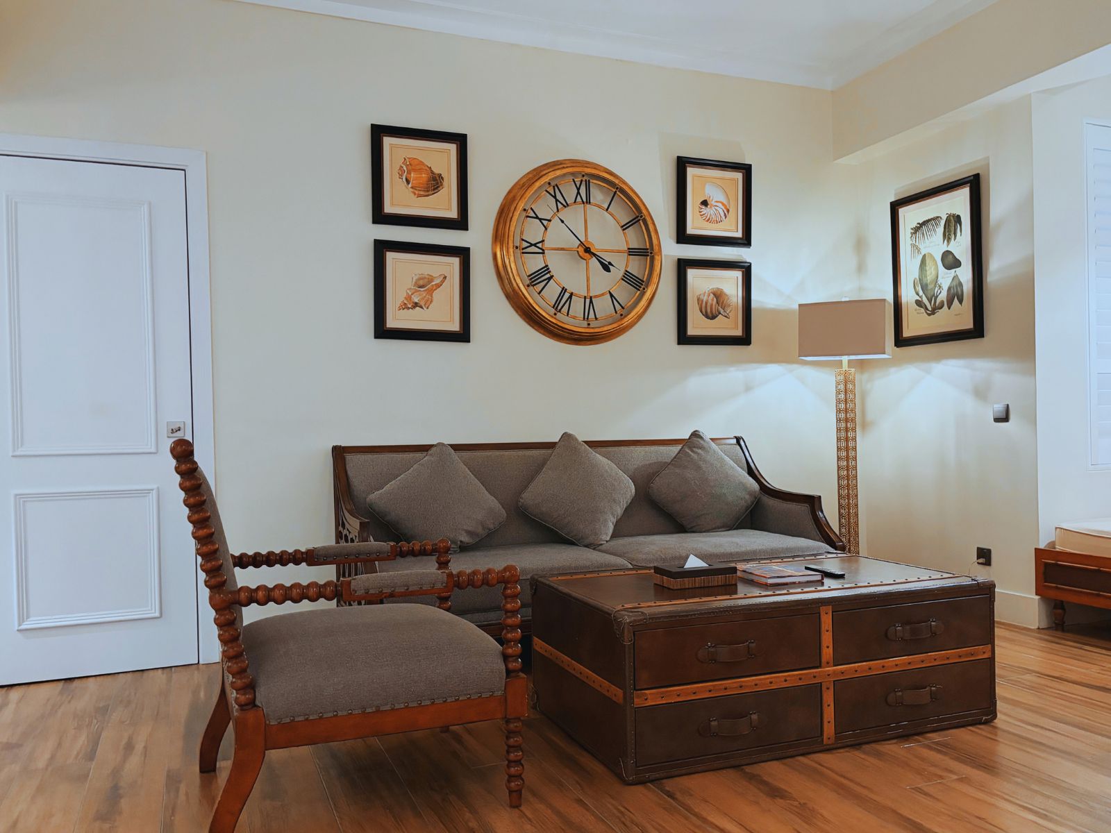 A living room with a sofa, a wooden chair, a large round wall clock, and framed pictures on the wall.