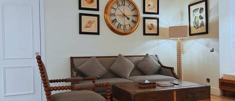 A living room with a sofa, a wooden chair, a large round wall clock, and framed pictures on the wall.