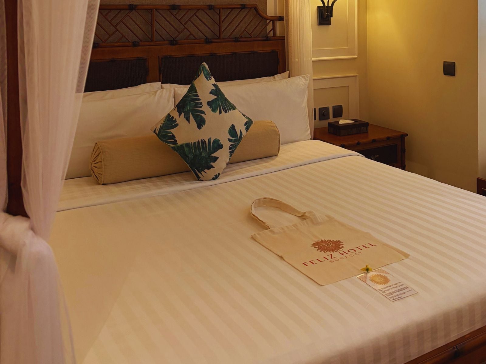 A neatly made bed with a striped bedspread, a pillow with a leaf pattern, and a hotel-branded tote bag on the bed.