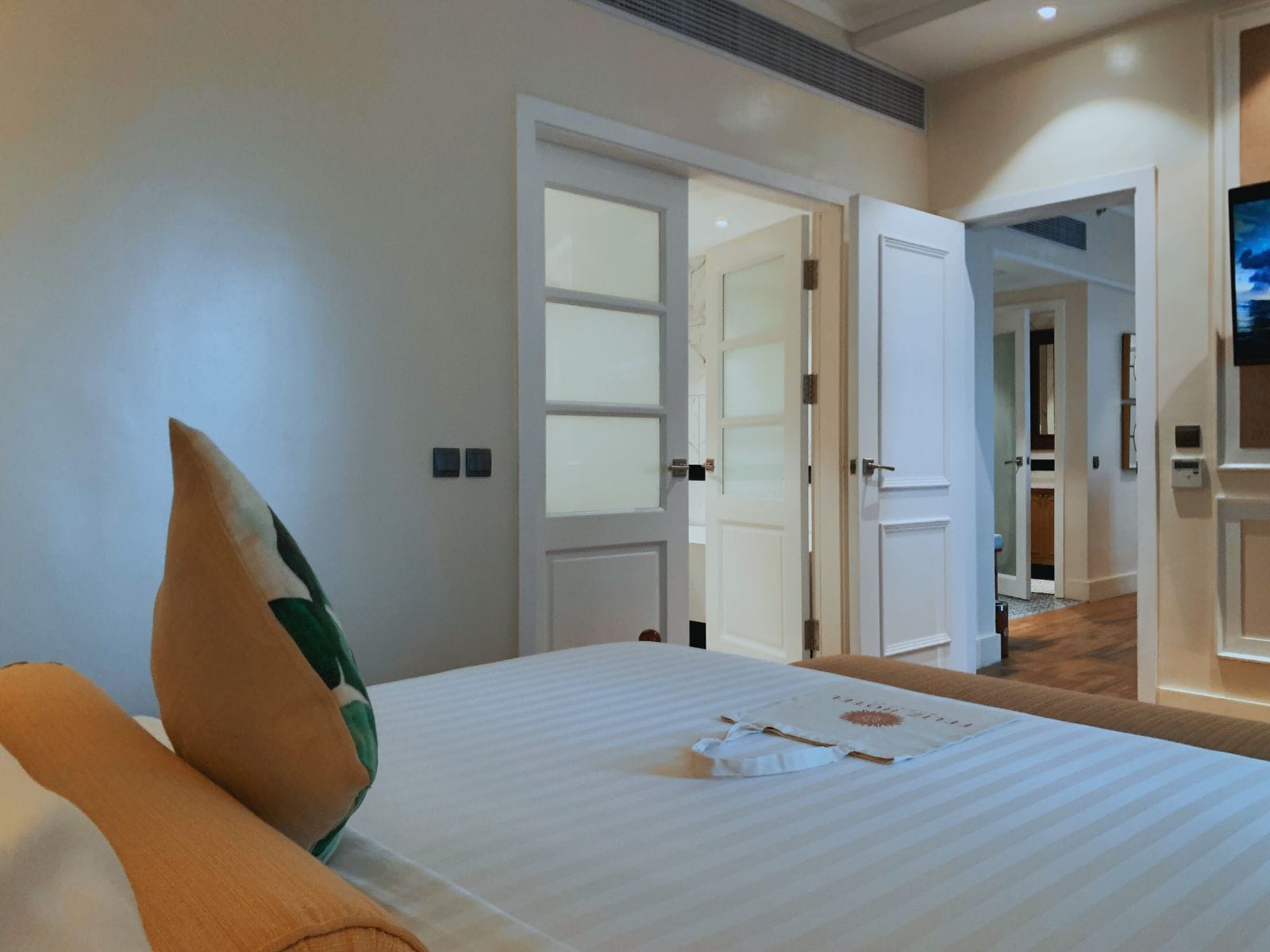 Una habitación de hotel con una cama, un almohadón y una puerta abierta que da a otra habitación.