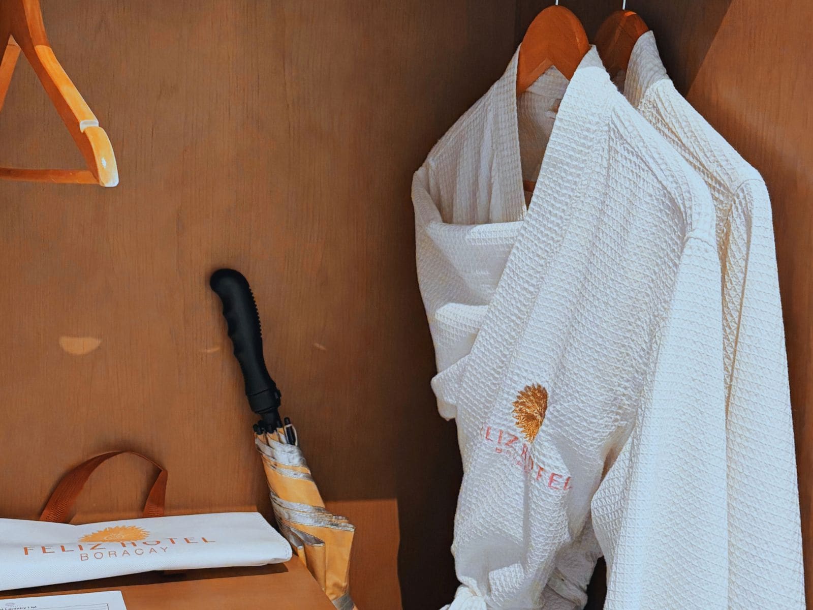 A wooden wall with a white towel hanging on a wooden hanger, a black-handled umbrella, and a white bag with 'FELIZ HOTEL BORACAY' text.