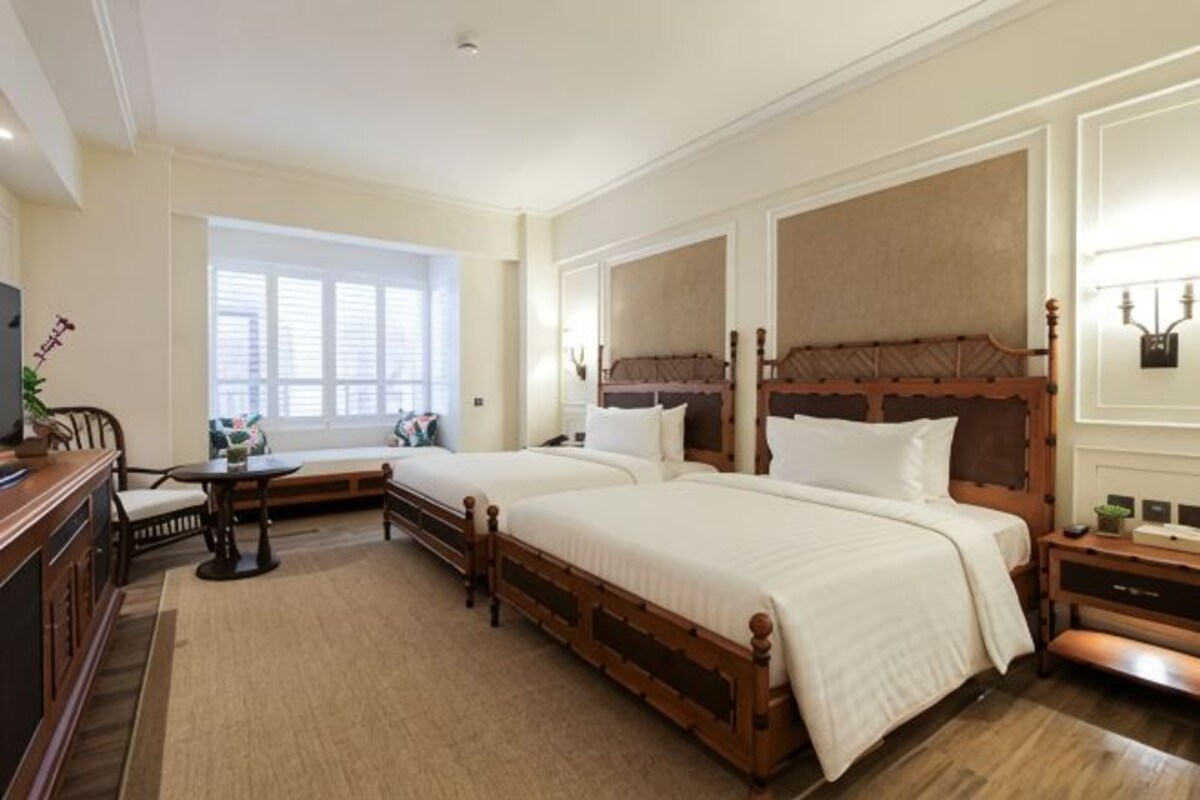 Habitación de hotel con dos camas, un escritorio, una silla, una ventana con persianas y una mesita de noche con lámpara.