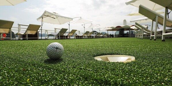 Una pelota de golf sobre césped artificial con un hoyo cerca, rodeado de sillones de jardín y sombrillas.