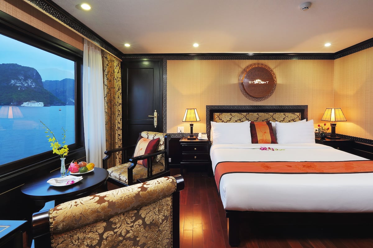 A hotel room with a bed, two bedside tables with lamps, a chair, a view of the sea, and a decorative wall piece.
