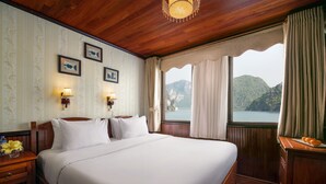 A neatly made bed with white linens, a wooden headboard, and two wall-mounted lamps. A window with curtains offers a view of a mountainous landscape. - Garden Bay Legend Cruise (Tuan Chau)