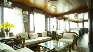 A spacious room with a wooden ceiling, large windows with curtains, a sofa, chairs, and a dining area with tables and chairs. - Garden Bay Legend Cruise (Tuan Chau)