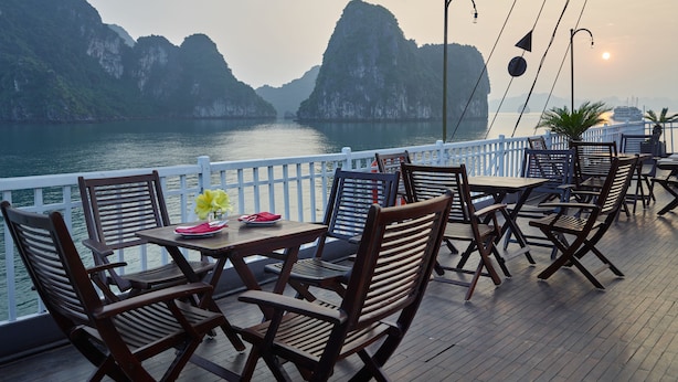 A deck with wooden chairs and tables, overlooking a body of water with mountains in the background. - Garden Bay Legend Cruise (Tuan Chau)