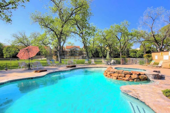 A swimming pool with lounge chairs and a red umbrella, surrounded by trees and a building in the background.