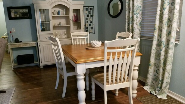 Un comedor con una mesa de madera y sillas blancas, un armario blanco con puertas de vidrio y un espejo en la pared.