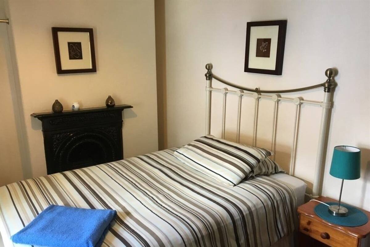 Een net opgemaakt bed met strepen, een blauwe handdoek, een houten nachtkastje met een lamp, en een open haard met decoratieve objecten.