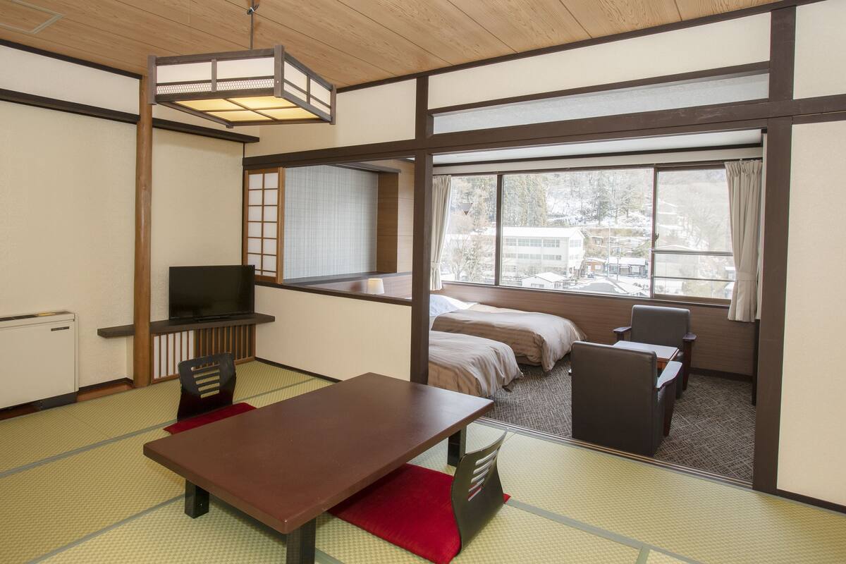 Une pièce de style japonais traditionnel, avec un sol recouvert de tatamis, une petite table en bois, un téléviseur et un lit donnant sur l’extérieur.
