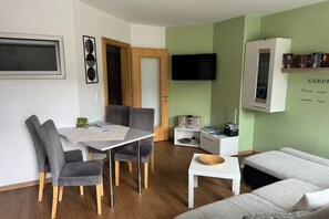 A modern living room with a dining area, a flat-screen TV mounted on the wall, a bookshelf, and a sofa.