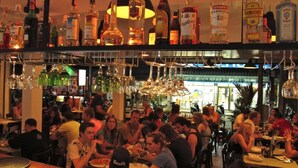 Un bar concurrido, con varias personas sentadas en mesas, una variedad de botellas de licor en estantes y cristalería colgante.