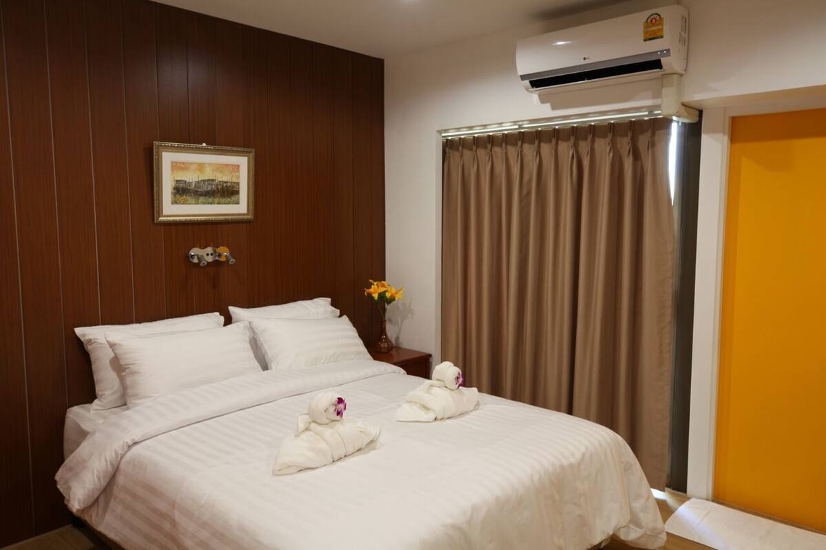 A hotel room with a bed, a wall-mounted air conditioner, a framed picture, a vase with flowers, and a curtain.