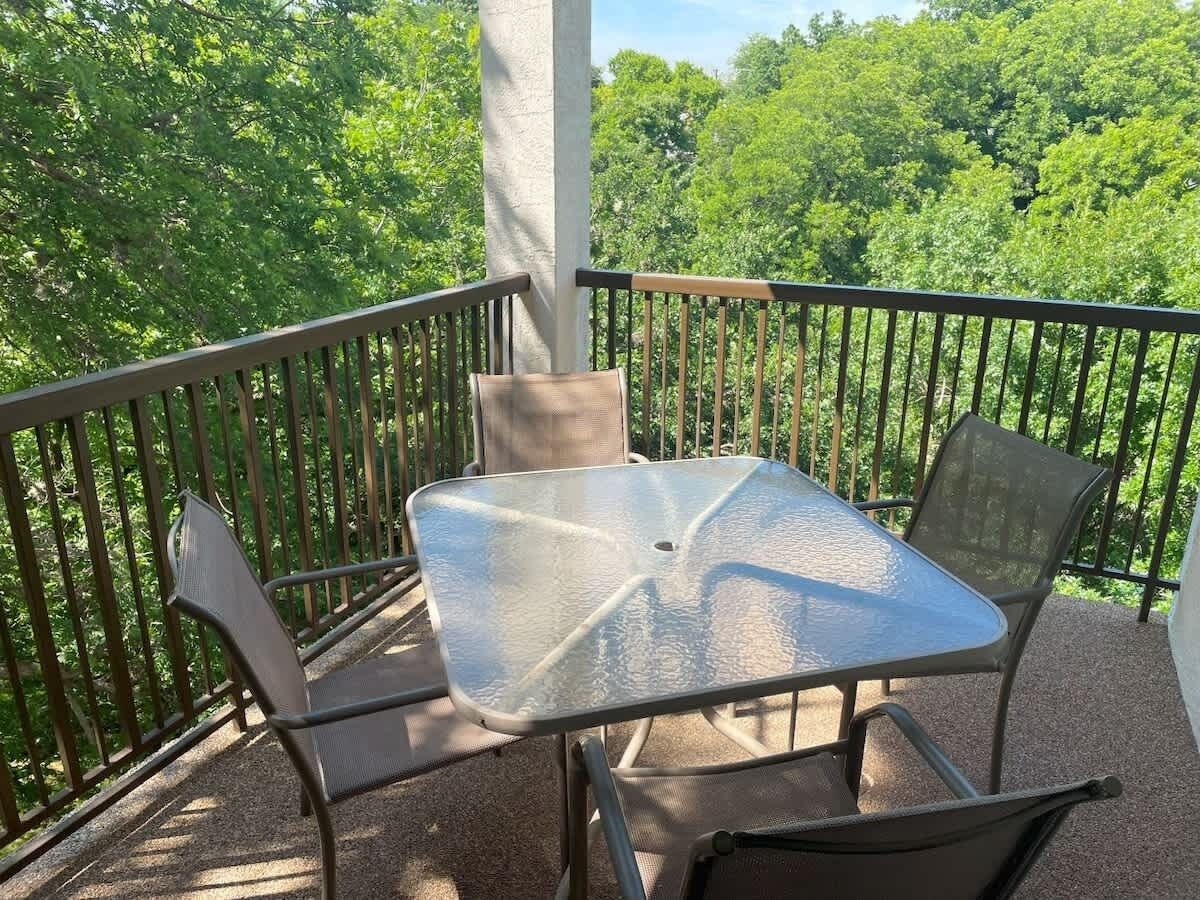 A balcony with a glass table and metal chairs, surrounded by a metal railing, overlooking a forested area.