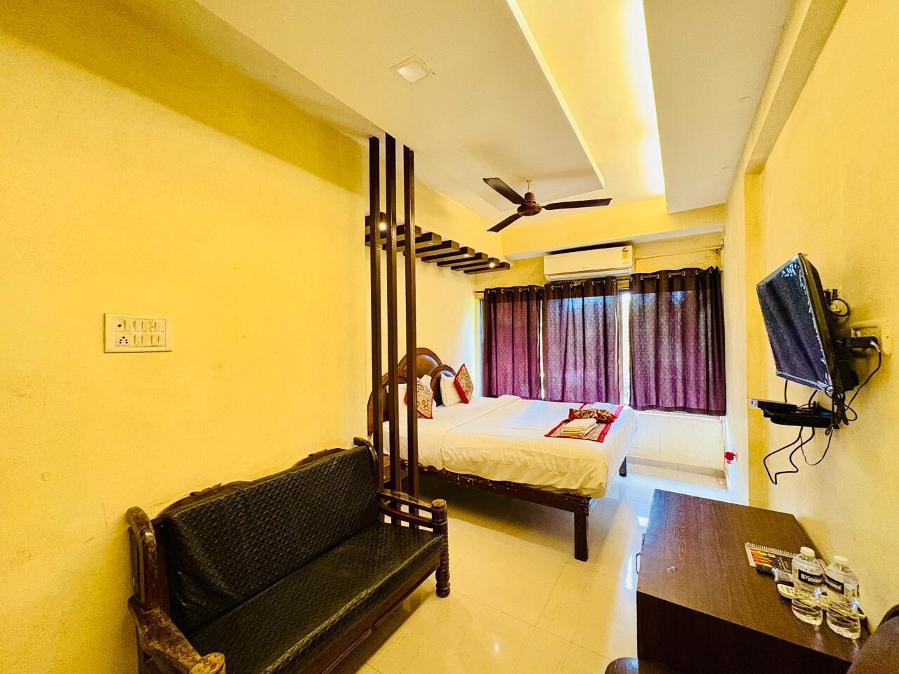 A hotel room with a bed, a sofa, a small table, a ceiling fan, and a window with curtains.