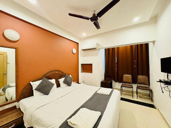 Habitación de hotel con una cama grande, ventilador de techo, equipo de aire acondicionado, televisión y una zona de estar con sillas.