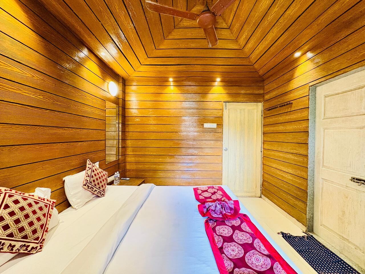 Ein Zimmer mit Holzwänden, einem Bett, Kissen und einer rot gemusterten Tagesdecke.