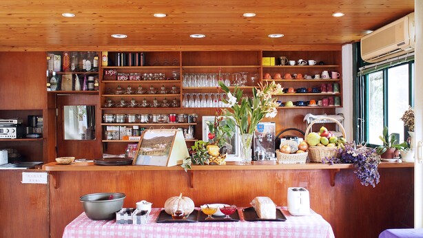 Interior de restaurante con estanterías de madera que exhiben cristalería y cerámica, una mesa con comida y bebidas, y una ventana con luz natural.
