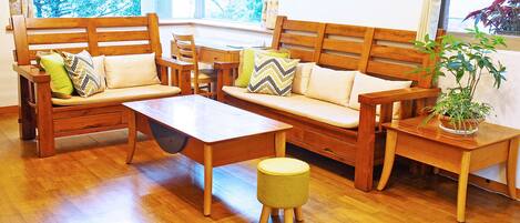A wooden sofa set with cushions, a small table, and a stool in a room with wooden flooring and a window.