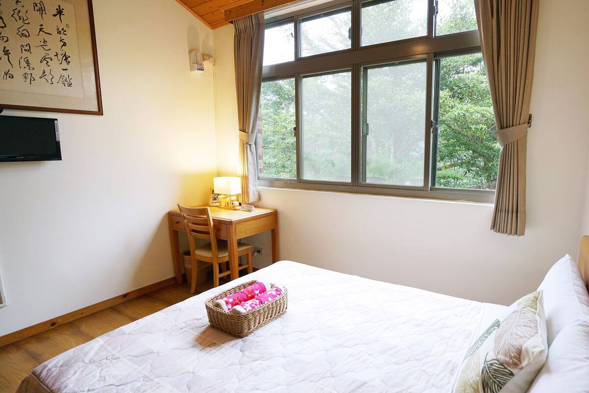 Quarto com cama, escrivaninha com cadeira, janela com cortinas e televisão fixada na parede.
