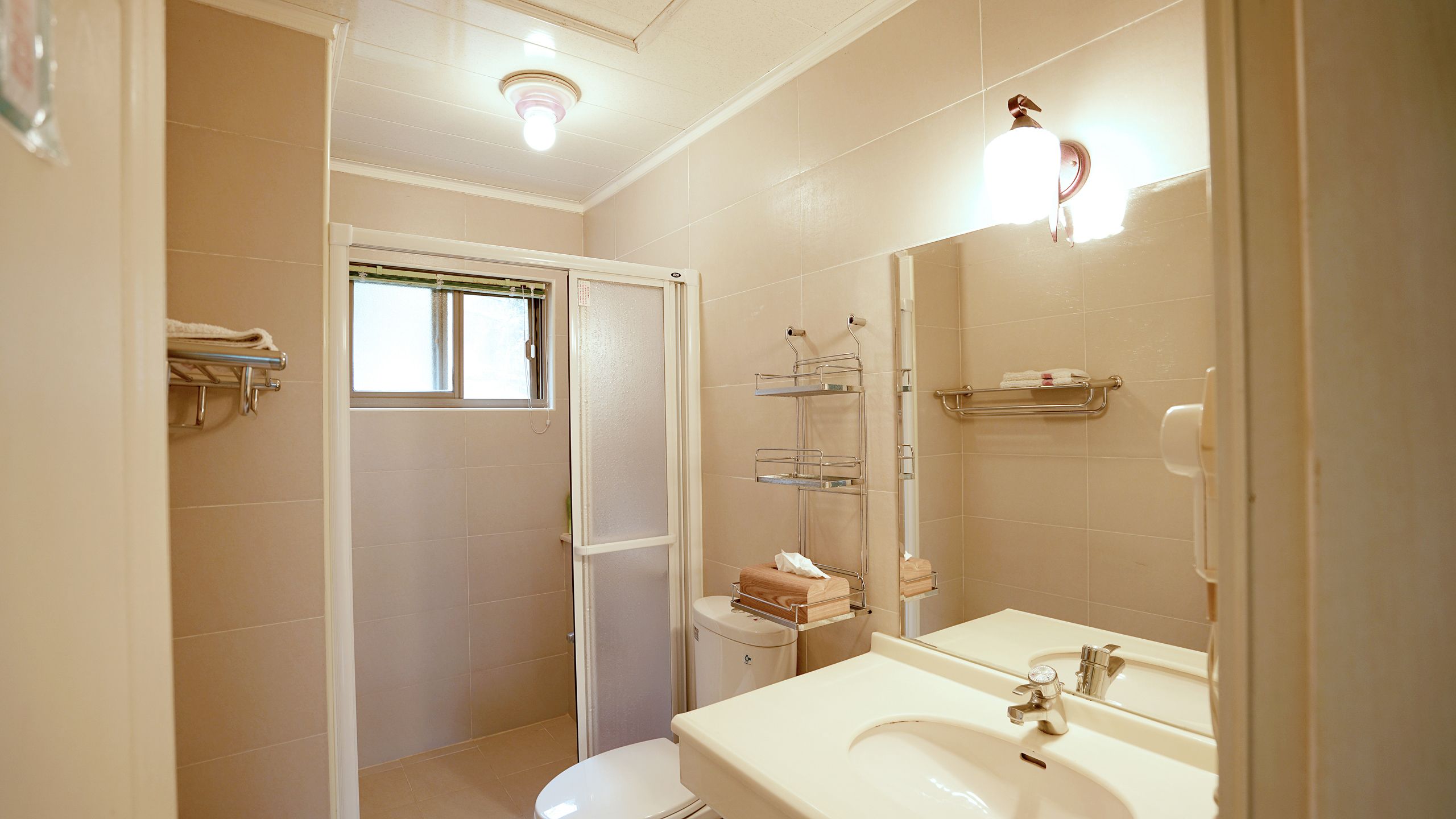 Une salle de bain comprenant des toilettes, un lavabo et un miroir. Il y a une fenêtre au-dessus des toilettes et un espace douche avec un porte-serviettes.
